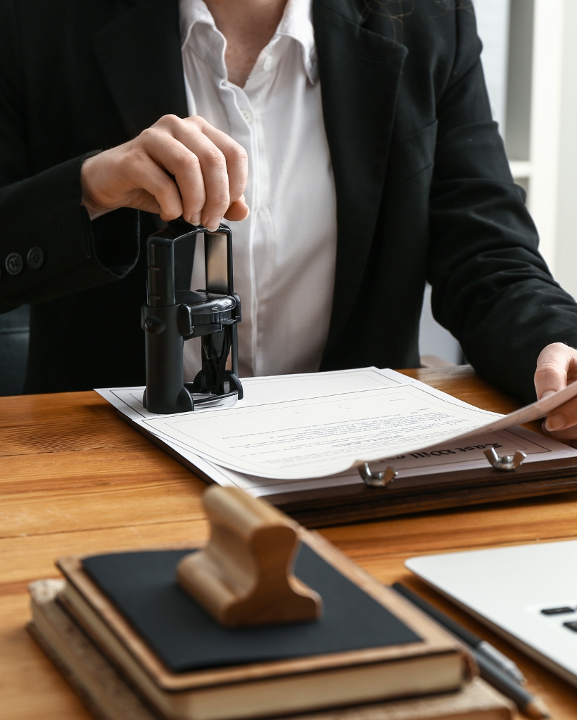 Notary in formal attire stamping a legal document at a desk with office supplies and a laptop, representing certified notary public services.