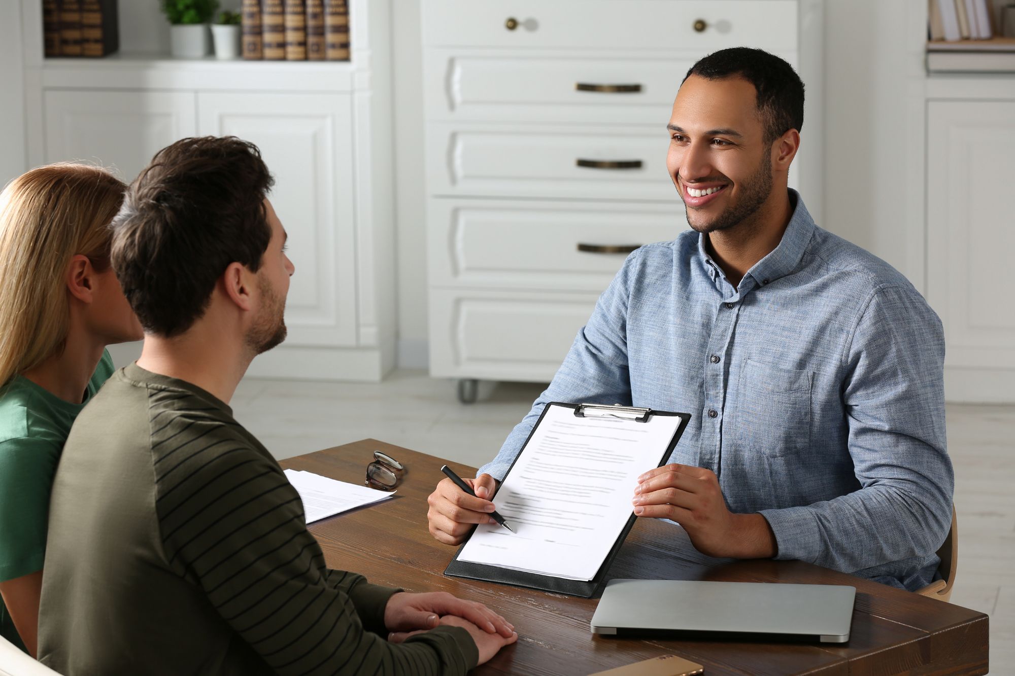 Smiling notary public reviewing documents with a couple at a desk during a consultation, holding a clipboard and pen ready for signatures.