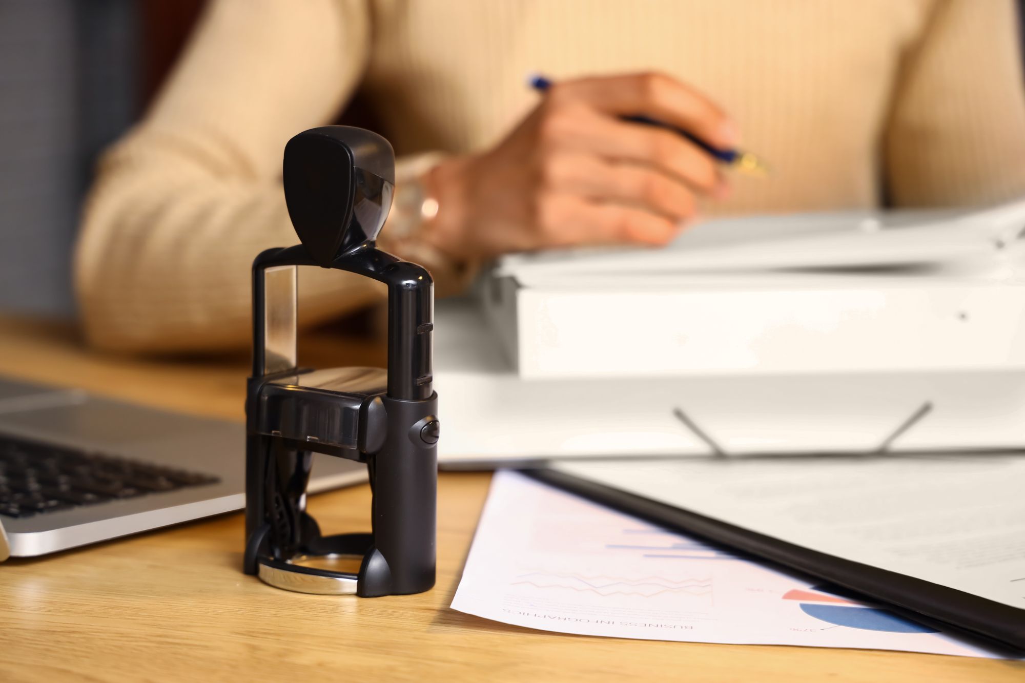 Modern self-inking notary stamp on a desk with business documents and a professional preparing paperwork in the background.