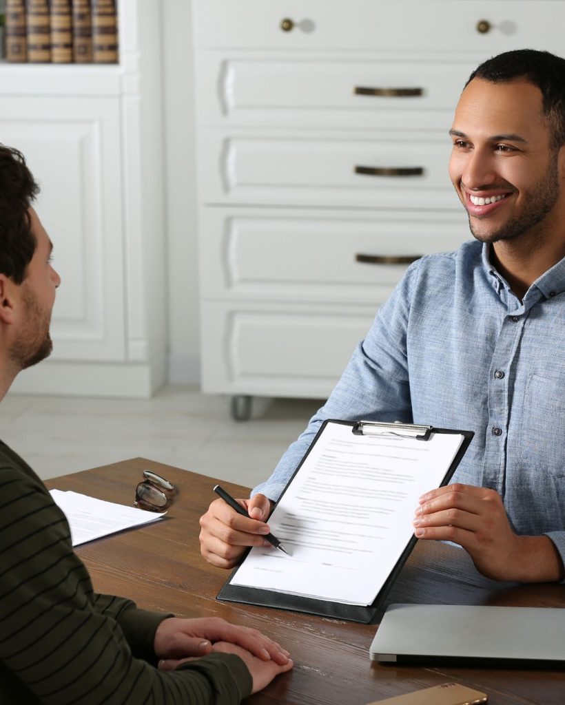 Smiling notary public reviewing documents with a couple at a desk during a consultation, holding a clipboard and pen ready for signatures.