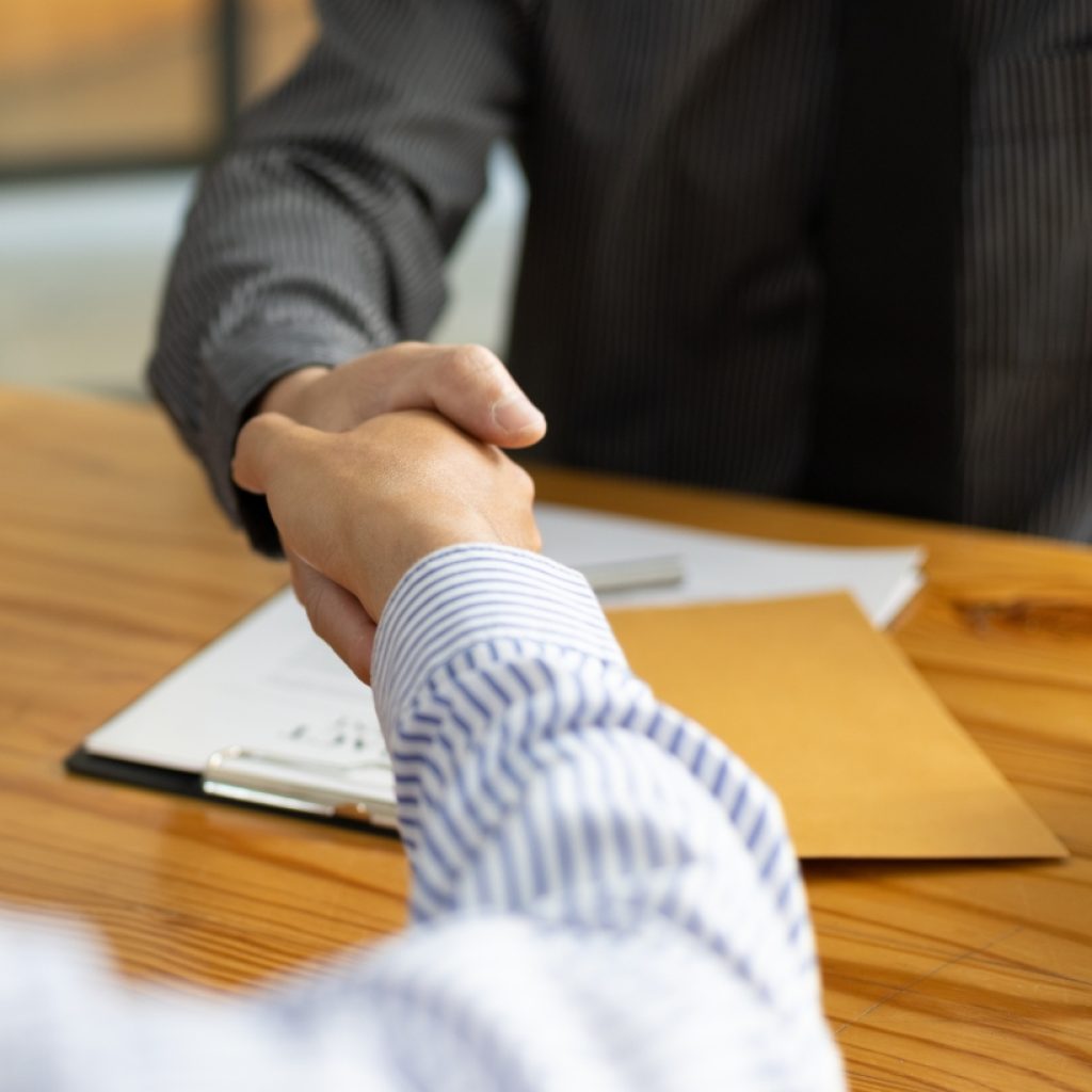 Two individuals shaking hands across a desk with legal documents and a manila envelope, symbolizing successful notary agreement or business deal.